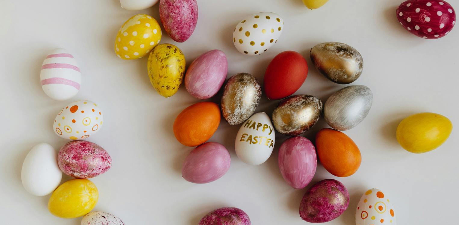 Flere fargerike påskeegg ligger strødd utover en hvit bakgrunn. På ett av dem står det "Happy easter".