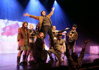 Ensemble av unge på scene i historiske klær; en mann står med åpne armer oppå lårene til to som kneler i en avslutningsposisjon.