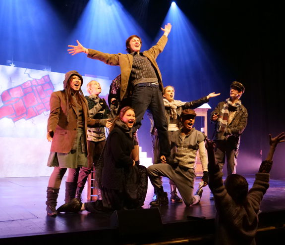 Ensemble av unge på scene i historiske klær; en mann står med åpne armer oppå lårene til to som kneler i en avslutningsposisjon.