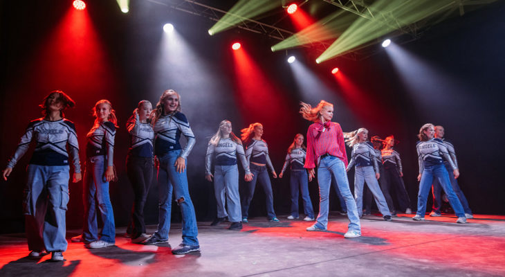 Ungdommer danser på en scene med grønt og rødt lys, kledd i hvite og blå sportsantrekk med teksten «Angels» på brystet.
