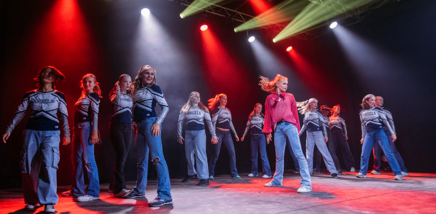 Ungdommer danser på en scene med grønt og rødt lys, kledd i hvite og blå sportsantrekk med teksten «Angels» på brystet.
