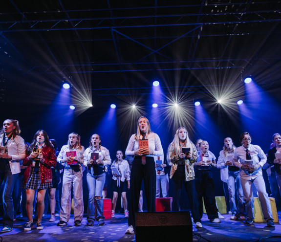 Ungdommer på scenen på Frilynt Norges sommerleir SplæshCamp, i ulike kostymer og med bøker i hendene, opplyst av hvite og blå scenelys.