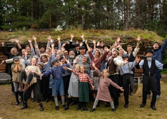 Ensemble fra utendørs teater foran en torvtak-hytte, kledd i historiske klær.