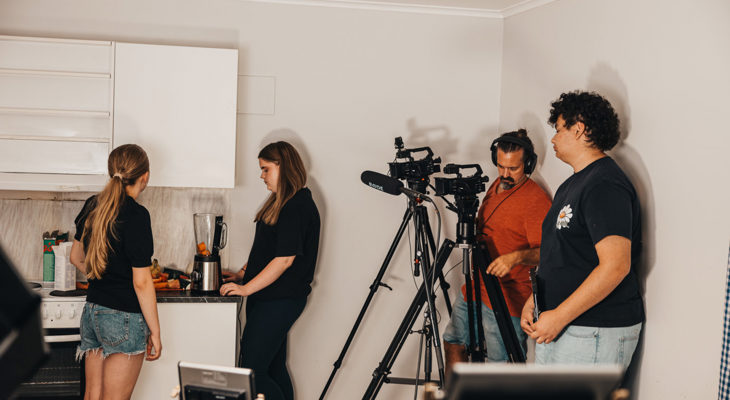 Tre ungdommer og en instruktør på kjøkkenet under et filmkurs; to kameraer filmer jentene som bruker en blender med klementiner.