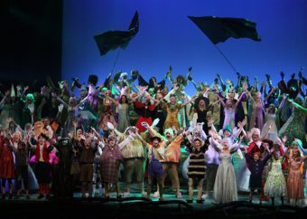 Ensemble fra Shrek The Musical i avslutningspositur med vaiende flagg i bakgrunnen.
