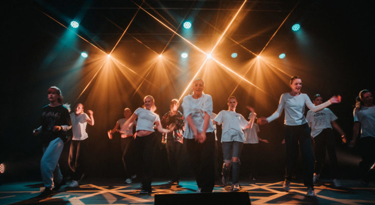 Ungdommer i hvite overdeler fremfører et dansenummer på scenen med sterkt gult og blått lys.