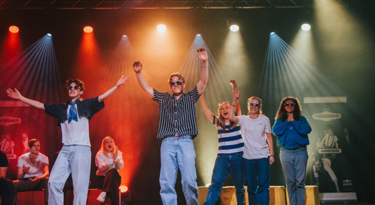 Ungdommer på scene med solbriller; to gutter i front klapper over hodet mens tre jenter står bak dem.