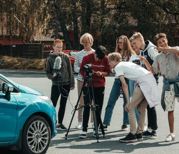 Ungdomsgruppe står foran et filmkamera som filmer en turkis bil; noen ser mot kameraet, andre mot fotografen.